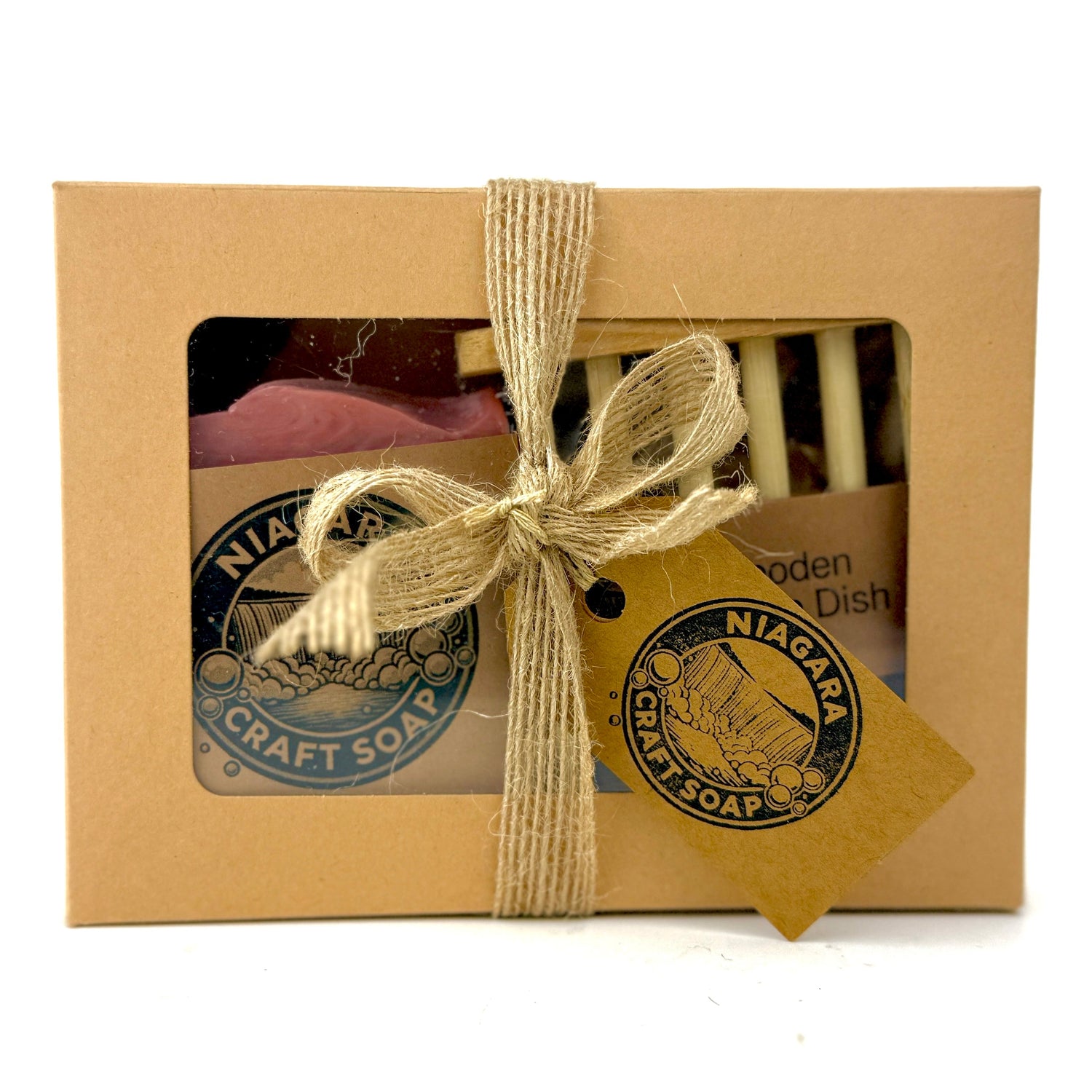 A Niagara Craft Soap Gift Set with bar soap and wooden soap dish in a small craft paper box seen through a cellophane window, wrapped in a jute fibre bow holding a craft paper tag with Niagara Craft Soap branding on one side