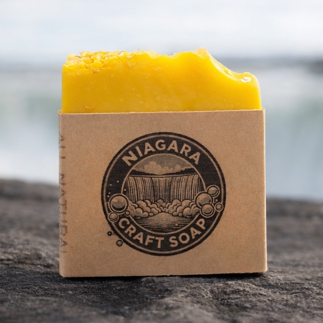 Bar of yellow soap on a brown cardboard block with 'Niagara Craft Soap' branding, set against a blurred natural background.