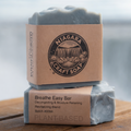 Two bars of soap with cardboard packaging on a wooden surface, with a blurred natural background.