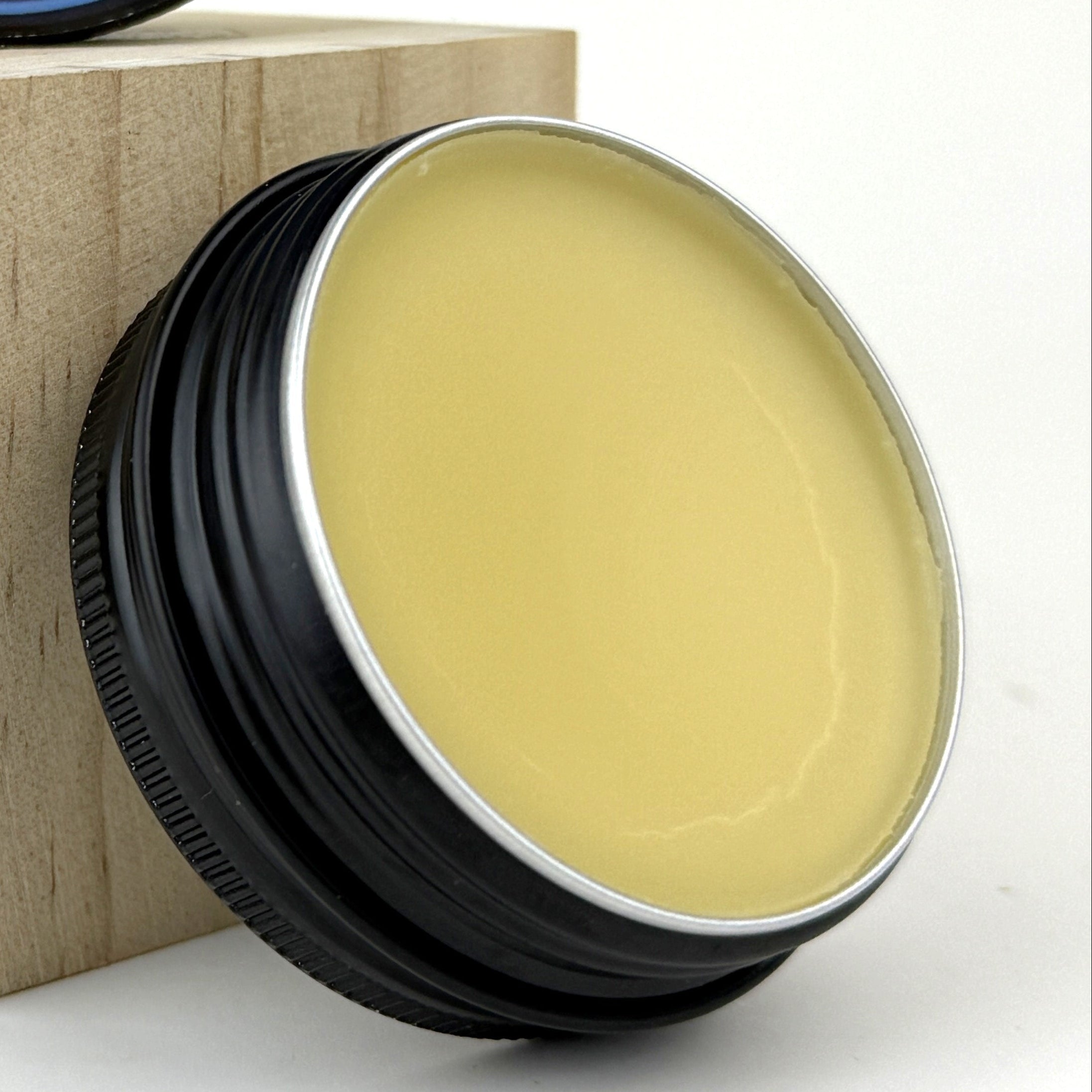 A twist cap jar of Niagara Craft Soap Tallow Whole Body Balm, with a thin ring of waxy craqueline around an other wise perfectly smooth, golden yellow balm, leaned along side a small wooden block.