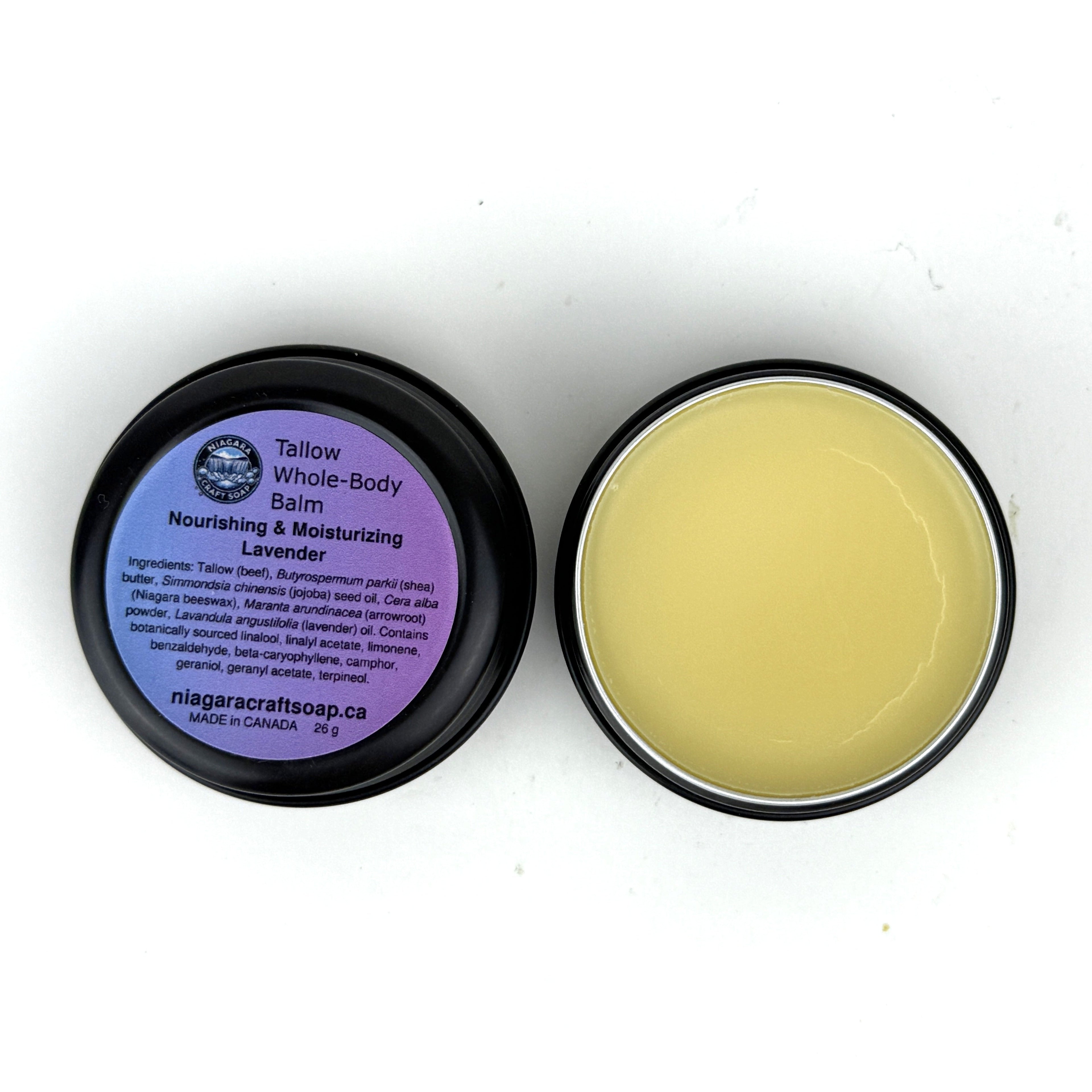 Two twist cap jars of Niagara Craft Soap Tallow Whole Body Balm, one open to demonstrate the golden yellow balm with, a thin wazy craqueline on an otherwise flawless surface, the other a tin turned with its branding including its ingredients facing upward, both sat on a white background with just a big of shadow around each tin. The tin itself is black with a matte to lustre like appearence.